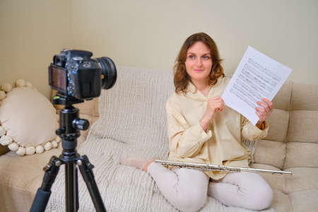 Woman musician with flute looking at video camera at home on sofa in living roomの写真素材