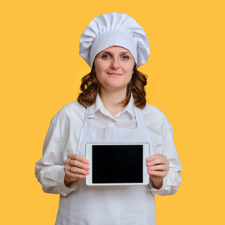 Happy woman chef showing ipad apple tablet screen on yellow studio background, mockup copy space - Moscow, Russia, October 20, 2021のeditorial素材