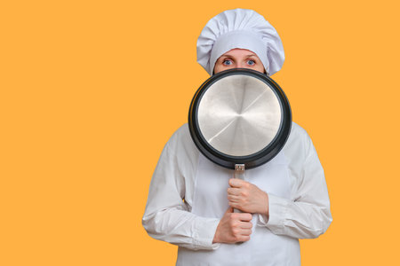 Frightened woman chef closes with a frying pan on yellow studio background, copy space