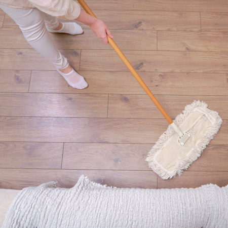 An adult woman mops a wooden laminate floor while cleaning her home living room.