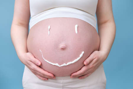 Cream in the form of a smile on the tummy of a pregnant woman, studio shot on a blue backgroundの写真素材
