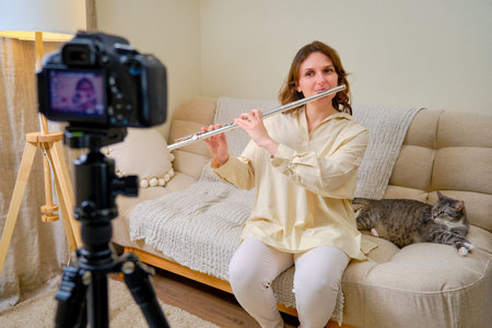 Happy woman musician with flute looking at video camera at home on sofa in living room