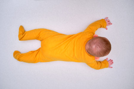 Infant baby lies on a play mat in yellow pajamas, top view. Toddler on the white floor full length,...