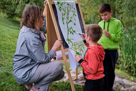 Mother teaches to paint with two boys pupils. Woman teacher artist paints with children on paper nature and trees by the riverの写真素材