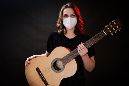 Female musician with a guitar in a medical face mask n95 on a dark studio background. Problems of...