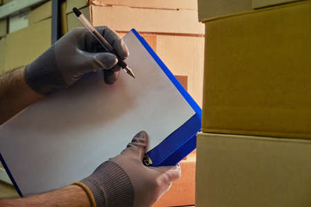 Men gloved hands write with a pen on a tablet with paper, checking a warehouse with goods in boxesの写真素材