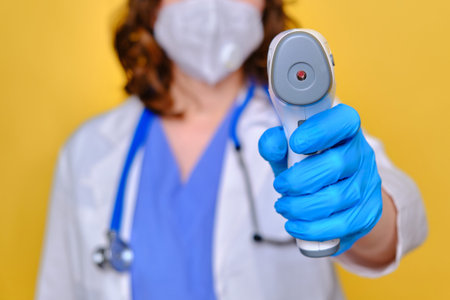 Woman doctor holds a infrared thermometer on a yellow background, close-up.