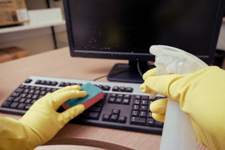Cleaning the office, a janitor in yellow gloves wipes keyboard and computer equipment with a rag.