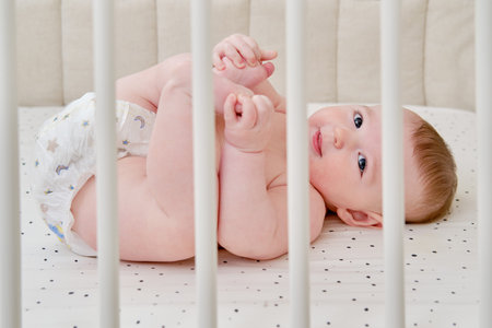 A happy infant baby with teething teeth in a diaper pulls his hand into his mouth. Funny child in a...