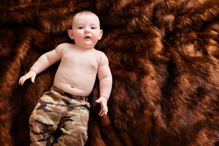 A funny happy baby boy is lying on a brown fur carpet in a military uniform. Smiling child with a...