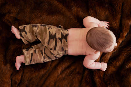 A funny happy baby boy is crawls on a brown fur carpet in a military uniform. Smiling child is lying on his stomach in khaki clothes, top viewの写真素材