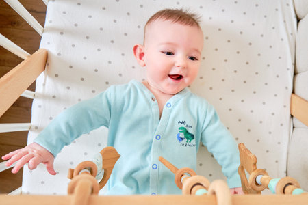 Happy infant baby is playing with wooden hanging toys lying in bed. Smiling funny child in turquoise clothes in a crib, aged six monthsの写真素材