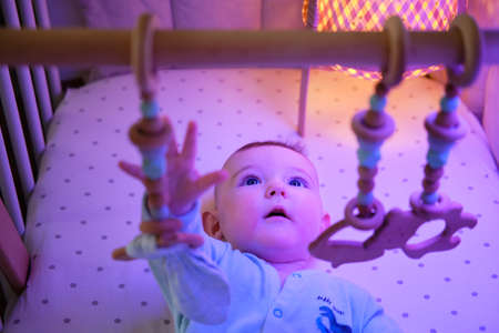 Curious infant baby is playing with wooden hanging toys in the evening in bed. Happy child in turquoise clothes in the light of a night lamp, age six monthsの写真素材