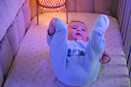Happy infant baby is playing with leg in the evening bed. Smiling child in turquoise clothes in the...