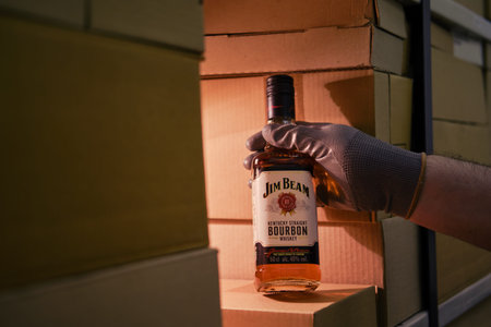 A man gloved hand holds a bottle of alcohol Jim Beam whiskey bourbon in a warehouse with boxes for delivery to customers and stores - Moscow, Russia, January 01, 2022のeditorial素材