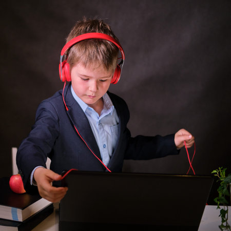 A happy boy in a school suit with headphones listens to music at the computer, copy space on a dark...