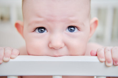 Baby gnaws at the edge crib during teething itching. Funny child scratching his teeth on the rail bed, age six monthsの写真素材