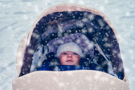 Stroller with a smiling toddler baby in a snowy winter park. Happy infant child in nature with falling snow, six to seven months oldの写真素材
