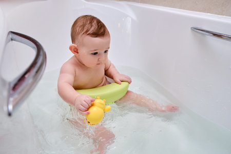 Toddler baby plays with a yellow duck toy while bathing. A happy infant child bathes in water while sitting on a chair in a white tub, six to seven months ageの写真素材