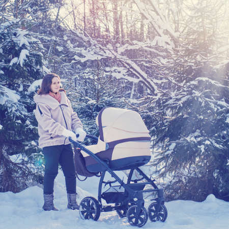 Woman mother with a stroller for a child in a winter park near snow-covered Christmas trees. Mom with a baby in a pram among snowy trees in the forestの写真素材