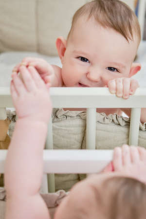 Happy baby is playing with her reflection in the mirror while standing in the crib. Funny lop-eared child pulls his hand to the mirror, six months oldの写真素材