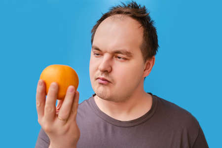 Portrait of a disheveled adult man with an orange fruit in his hand, blue studio backgroundの写真素材