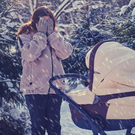 Woman mother crying at pram with baby in winter park. Sad mother with a baby in a stroller among...