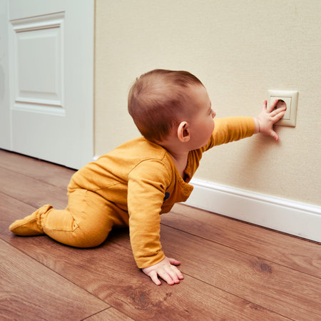 Baby toddler reaches into the electrical outlet on the home wall with his hand. Danger and...