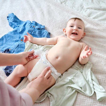 A mother woman puts on a diaper for a baby boy. Mom dresses a happy child in clothes on the bed.の写真素材
