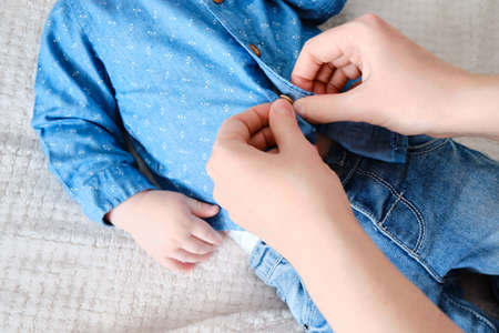 A mother woman wears a blue shirt to a baby boy. Mom dresses a child in clothes on the bed.の写真素材