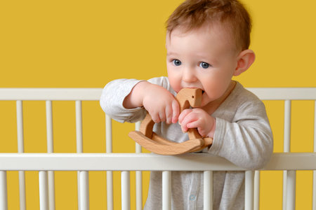Sad toddler baby boy in the crib gnawing a wooden toy, yellow studio background. A tired child in pajamasの写真素材