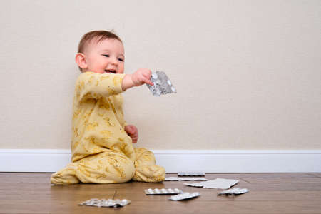 Toddler baby boy plays with medicines in a package while sitting on the floor. Child holding medical pills and drug in his handsの写真素材