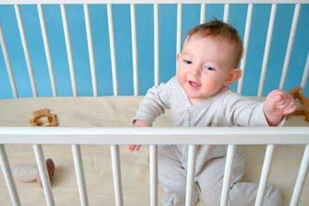 Happy infant baby boy is sitting in a crib, studio blue background. Smiling child in white pajamas, copy spaceの写真素材