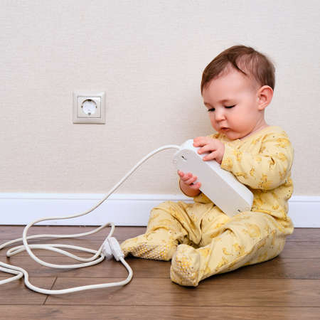 Toddler baby boy plays with electric wires while sitting on the floor. Child holding an extension cord with electrical socketsの写真素材
