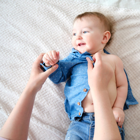 A mother woman wears a blue shirt to a baby boy. Mom dresses a happy child in clothes on the bed.