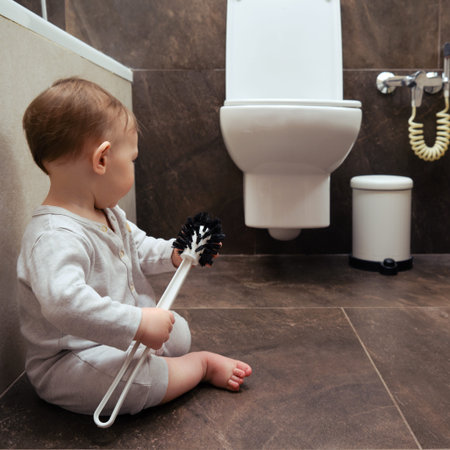 Toddler baby boy is playing in the toilet room with a brush. Child plays on the brown floor in the...