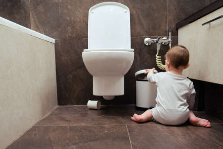 Toddler baby boy is playing in the toilet room with a trash can. Child plays on a brown floor in a beige bathroom with a trash can lidの写真素材