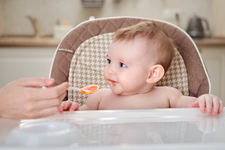 Mother feeding toddler baby from a spoon on a high chair for children, home kitchen background....