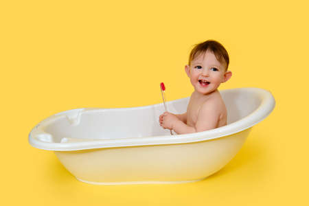 Happy baby toddler boy with a toothbrush in a white bathtub on a studio yellow background. A smiling child at the age of one year, copy spaceの写真素材