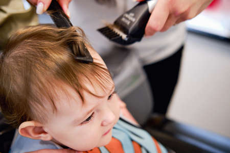 The hairdresser cuts the toddler baby boy in the barbershop. Child first haircut. Kid age one year (12 months)の写真素材