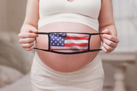 Medical face mask with the US flag in the hands of a pregnant woman, home living roomの写真素材