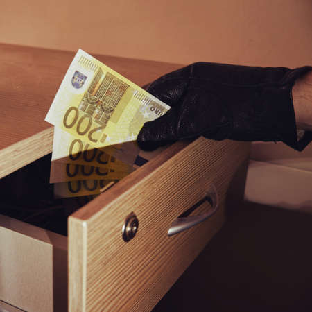 Man hand in a black glove takes money out of an office desk drawer in the night light of a flashlightの写真素材