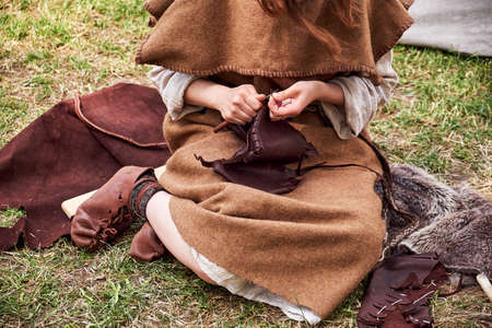 A woman in old Roman clothes sews leather sitting on the grass. Reconstruction of the events of the Roman Empireの写真素材