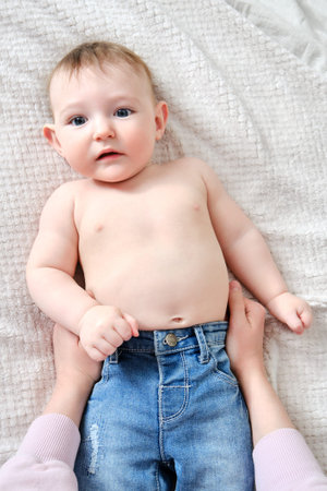 A mother woman puts on jeans pants for a baby boy. Mom dresses a happy child in clothes on the bed.