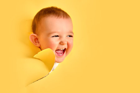 A happy child looks out of a hole in the studio yellow background. Smiling baby boy peeks through a...
