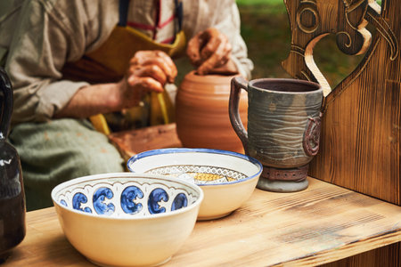 A man in old medieval Byzantine clothes sits behind a vintage potter's wheel and makes dishes out of...