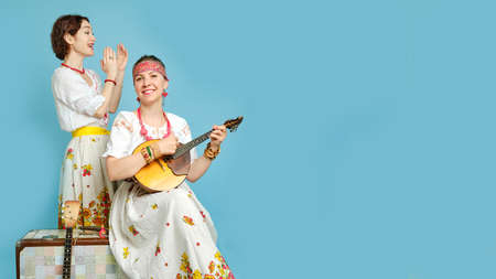 Women musicians in Russian folk dresses with musical instruments on a blue studio background. Happy artists from Russia in white national clothes with stringed musical instrumentsの写真素材