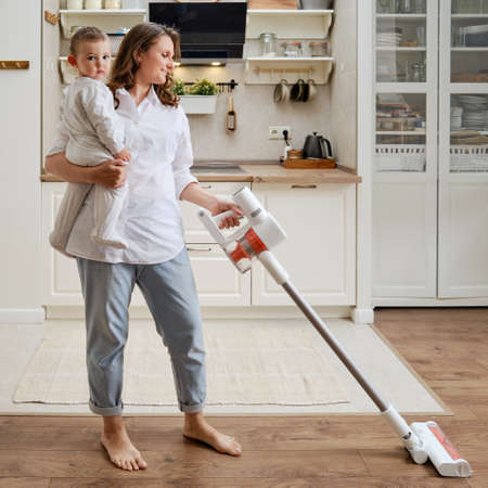 Happy woman holds baby and wireless portable vacuum cleaner. A smiling woman with a child in her arms cleans the floor in an apartment with a vacuum cleanerの写真素材