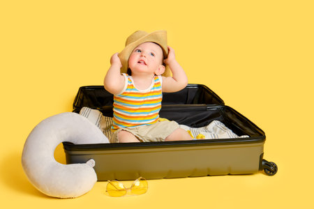 Baby toddler boy sits inside a suitcase with clothes, studio yellow background. A child plays in a big bag while packing luggage for a vacation trip, copy space. Kid age one year (12 months)の写真素材
