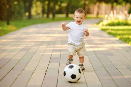 Happy toddler baby boy is playing walking behind a soccer ball on a stone path, first steps. Smiling child in white clothes walks with a ball, age one yearの写真素材